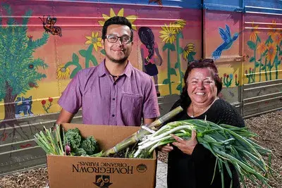 A man and a woman smiling while holding fresh vegetables, standing in front of a colorful mural with plants and animals.